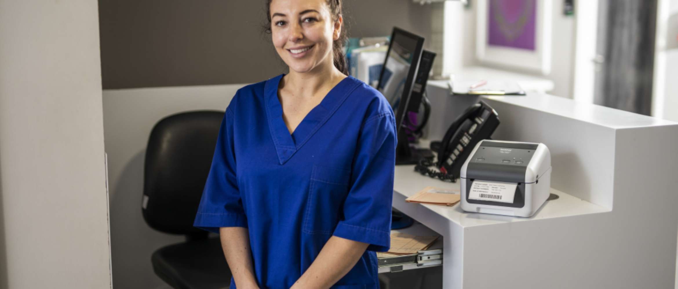 A female doctor wearing blue hospital scrubs is standing in front of a medical centre's back office containing a computer and Brother labelling printer in this healthcare scene