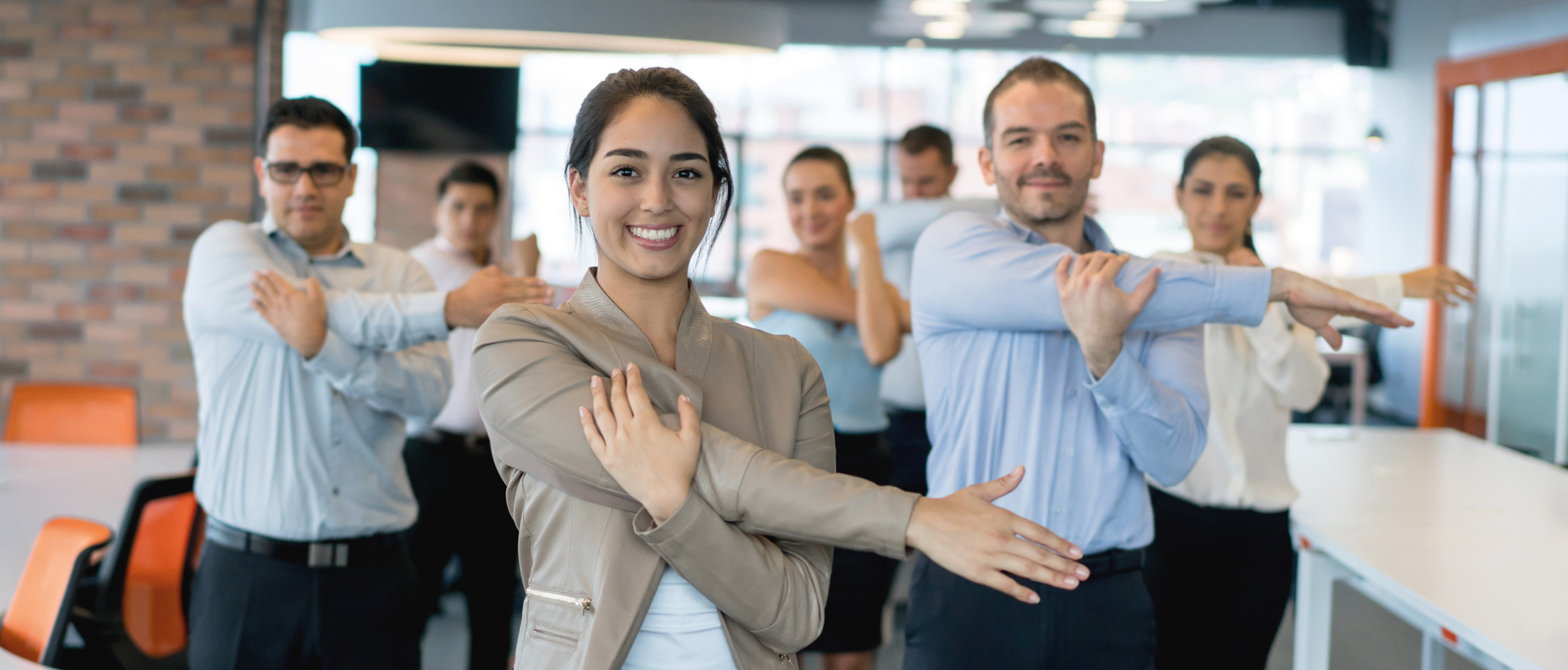 Seven office workers in casual business attire - four male and three female - are exercising with arm stretches in this scene on staff wellbeing