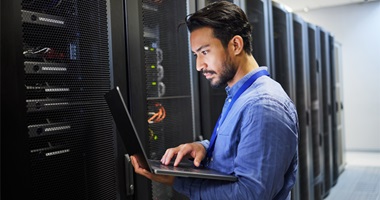 A person standing in front of a server rack, using a laptop. 