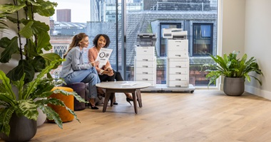 Two people seated at a small round table in a modern office with two business printers in the background