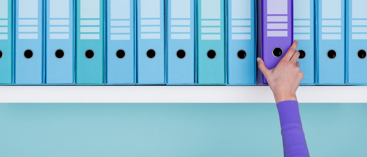 A shelf full of similar looking teal blue-green lever arch files has one purple file folder standing out. It is being picked up by an office staff member wearing a matching purple item of clothing.