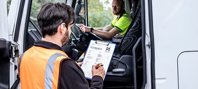 En man i en orange väst fyller i ett dokument medan han pratar med en förare som sitter i lastbilen